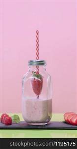 Tasty looking milkshake decorated with a straw piercing a strawberry inside a glass jar bottle on a dark plate with raspberries and strawberry slices on a pink background. Healthy and vegetarian food concept.. Tasty looking strawberry milkshake in a glass bottle jar