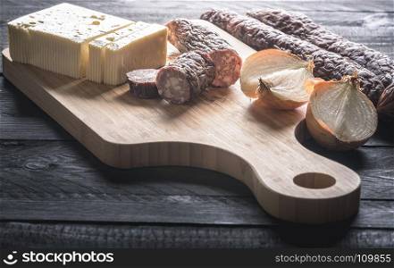 Tasty meal with homemade smoked sausages from beef and pork, fresh cheese and onion, displayed on a wooden cutting board, on a rustic black table.