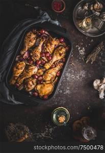 Tasty roasted chicken drumsticks with red onion and grapes in cast iron casserole pan on dark rustic background, top view