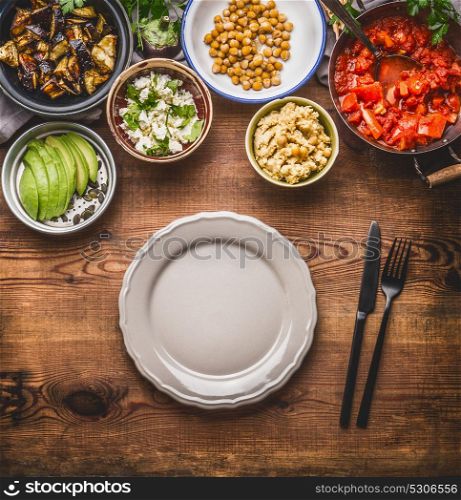 Tasty vegetarian ingredients for healthy meal in bowls: chick peas puree, roasted vegetables , red paprika stew, avocado and seeds served with empty plate with cutlery Clean eating food concept
