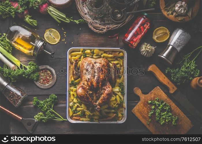 Tasty whole roasted chicken on baking tray with potatoes served on dark rustic kitchen table with cooking ingredients , canned and fresh vegetables , olives oil, lemon, herbs and spices. Top view.