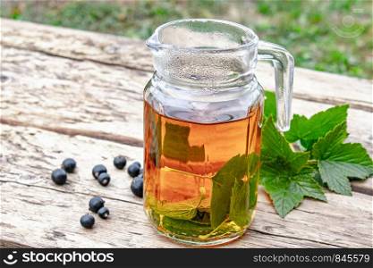 Tea in a glass teapot with green leaves of black currant on a wooden table against the background of green grass. Close-up.. Tea in a glass teapot with green leaves of black currant on a wooden table against the background of green grass.