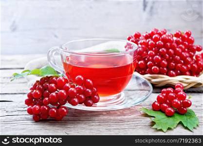 Tea with viburnum in a glass cup, green leaves, wicker plate with berries and a burlap napkin on wooden board background