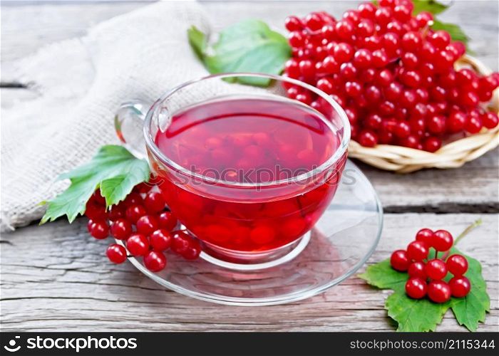 Tea with viburnum in a glass cup, wicker plate with berries, green leaves and burlap napkin on wooden board background