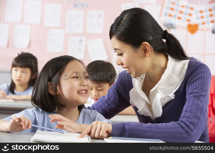 Teacher Helping Student Working At Desk In Chinese School Classroom