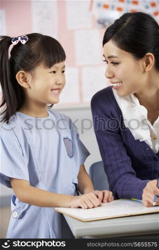 Teacher Helping Student Working At Desk In Chinese School Classroom