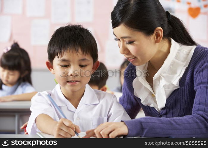 Teacher Helping Student Working At Desk In Chinese School Classroom