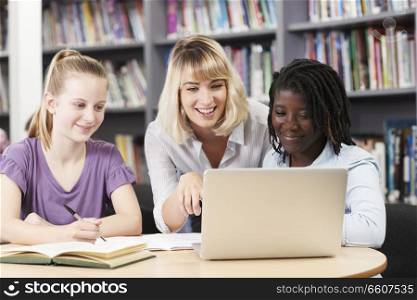 Teacher Helping Two Female High School Students Working At Laptop In Library