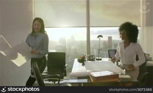 Team of two female architects standing in modern office with building plan. The women talk and review a housing project smiling and pointing on paper. Steadicam shot with lens flare
