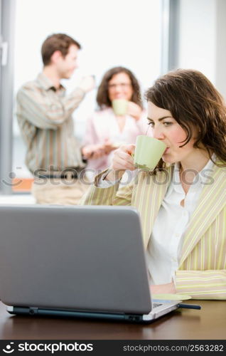 Team of young successful businesspeople talking and drinking coffee at office, businesswoman working on laptop computer in front.