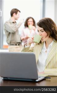 Team of young successful businesspeople talking and drinking coffee at office, businesswoman working on laptop computer in front.