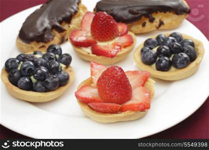 Teatime treats of homemade tarts filled with confectioner&rsquo;s custard and topped with fresh strawberries or blueberries, and chocolate eclairs. Normal focus.