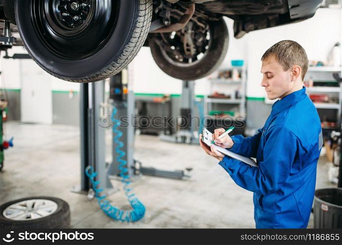 Technician with notebook fills the check list, car on the lift, fixing the problems. Automobile service, vehicle maintenance