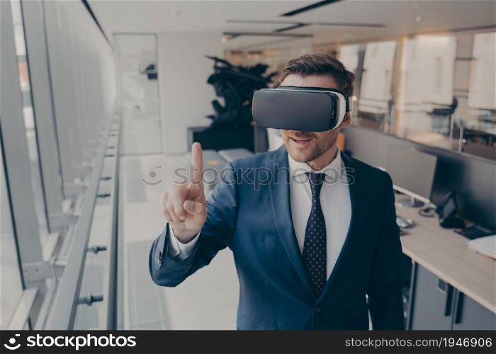 Technologies of future. Young businessman in elegant suit using VR headset, touching exploring virtual reality at work, trying to touch something with forefinger in front of him, standing in office. Young businessman in elegant suit using VR headset, touching exploring virtual reality at work