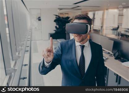 Technologies of future. Young businessman in elegant suit using VR headset, touching exploring virtual reality at work, trying to touch something with forefinger in front of him, standing in office. Young businessman in elegant suit using VR headset, touching exploring virtual reality at work