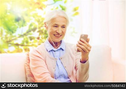 technology, age and people concept - happy senior woman with smartphone and earphones listening to music at home over window with green natural background. senior woman with smartphone and earphones at home. senior woman with smartphone and earphones at home