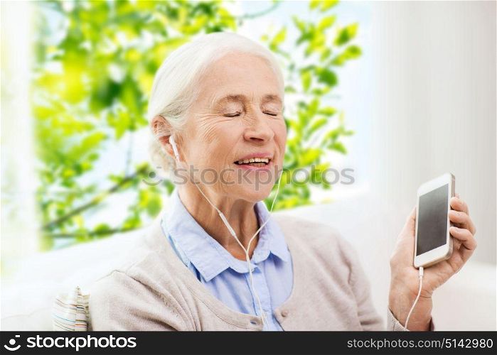 technology, age and people concept - happy senior woman with smartphone and earphones listening to music at home over window and green natural background. senior woman with smartphone and earphones at home