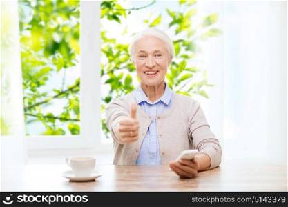 technology, age and people concept - happy senior woman with smartphone and coffee sitting at table and showing thumbs up at home over window and green natural background. senior woman with smartphone showing thumbs up