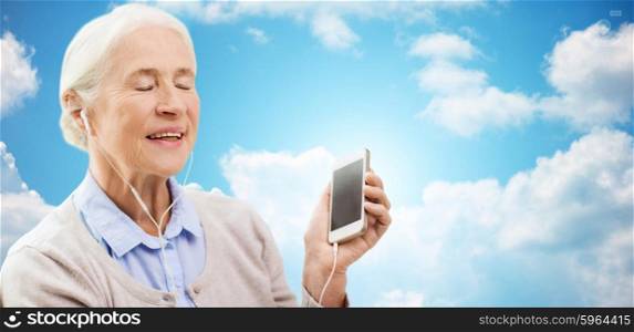 technology, age and people concept - happy senior woman with smartphone and earphones listening to music over blue sky and clouds background