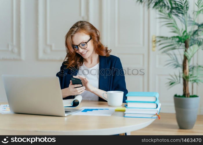 Technology and communication concept. Red haired young female checks email box on modern cell phone, has pleasant smile, studies documents, owns big corporation, uses laptop computer for work