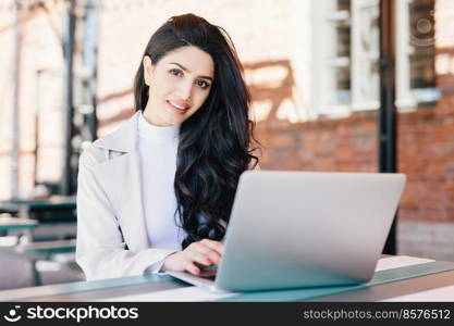 Technology and communication concept. Successful European businesswoman with beautiful appearance working at a cafe on laptop computer having smiling expression looking in camera while sitting outdoor