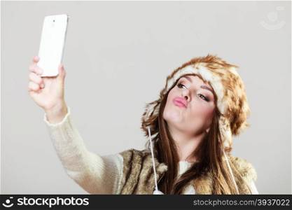 Technology and fun concept. Beauty young happy woman in warm winter clothes taking selfie photo self picture using phone in studio on gray.
