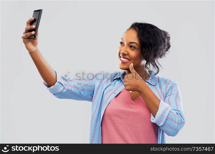 technology and gesture people concept - happy african american woman taking selfie by smartphone and showing thumbs up over grey background. african american woman taking selfie by smartphone