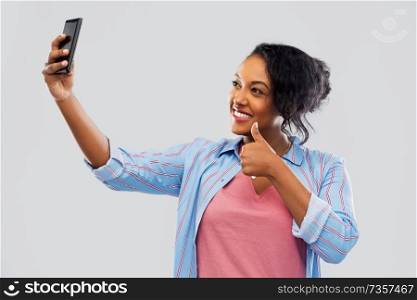 technology and gesture people concept - happy african american woman taking selfie by smartphone and showing thumbs up over grey background. african american woman taking selfie by smartphone