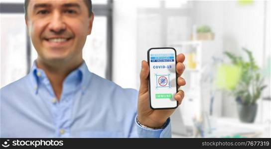 technology and health care concept - close up of smiling man holding and showing smartphone with international certificate of vaccination on screen over office background. man with certificate of vaccination on smartphone