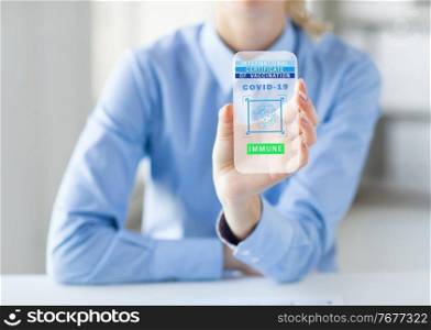 technology and health care concept - close up of woman showing transparent smartphone with international certificate of vaccination or virtual immunity passport on screen at office. hand with certificate of vaccination on smartphone