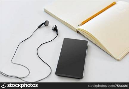 technology and objects concept - earphones, smartphone and notebook with pencil on white background. earphones, smartphone and notebook with pencil