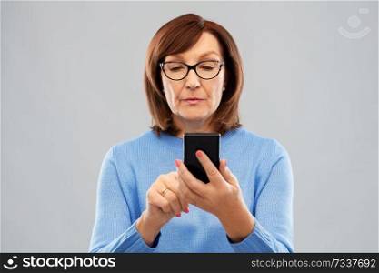 technology and old people concept - senior woman in glasses using smartphone over grey background. senior woman in glasses using smartphone