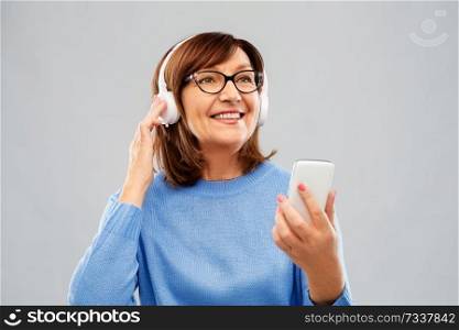 technology and old people concept - smiling senior woman in glasses and headphones listening music on smartphone over grey background. senior woman in headphones listening music on cell