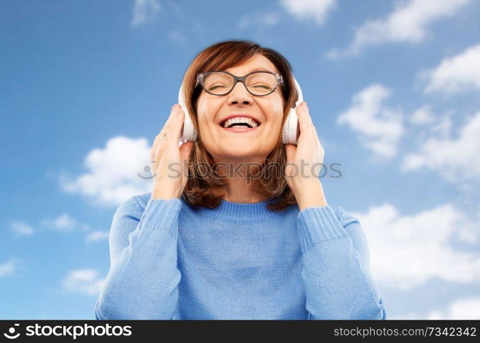 technology and old people concept - smiling senior woman in glasses and headphones listening to music over blue sky and clouds background. senior woman in headphones listening to music