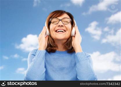 technology and old people concept - smiling senior woman in glasses and headphones listening to music over blue sky and clouds background. senior woman in headphones listening to music