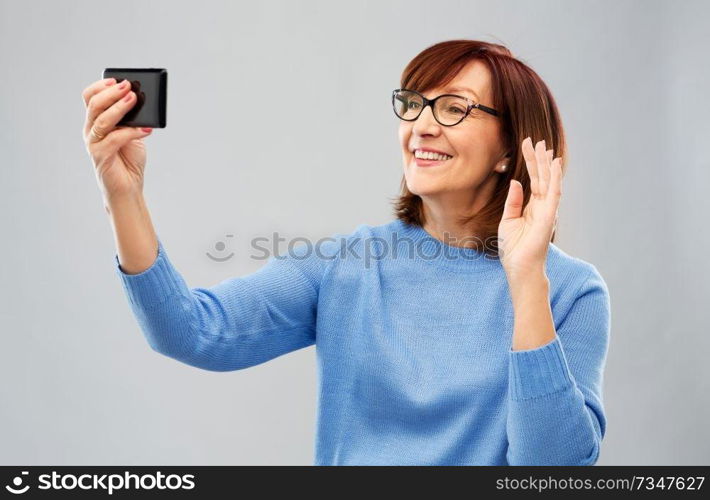 technology and old people concept - smiling senior woman in glasses taking selfie or having video call on smartphone over grey background. senior woman having video call on smartphone