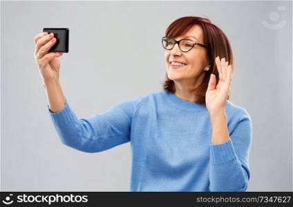 technology and old people concept - smiling senior woman in glasses taking selfie or having video call on smartphone over grey background. senior woman having video call on smartphone