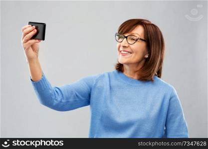 technology and old people concept - smiling senior woman in glasses taking selfie by smartphone over grey background. senior woman taking selfie by smartphone
