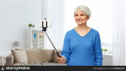 technology and old people concept - smiling senior woman taking picture by smartphone on selfie stick over home living room background. smiling senior woman taking selfie by smartphone