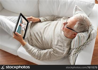 technology and online communication concept - senior man with tablet pc computer lying on sofa at home and having video call with woman. old man with tablet pc has video call with woman
