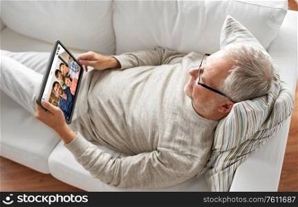 technology and online communication concept - senior man with tablet pc computer lying on sofa at home and having video call with family. senoir man having video call with family