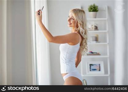 technology and people concept - beautiful young woman in white underwear taking selfie by smartphone at home in morning. woman taking selfie by smartphone in morning