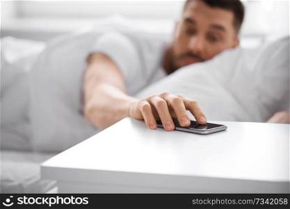 technology and people concept - close up of sleepy young man reaching for smartphone on bedside table from bed at home in morning. sleepy young man reaching for smartphone in bed