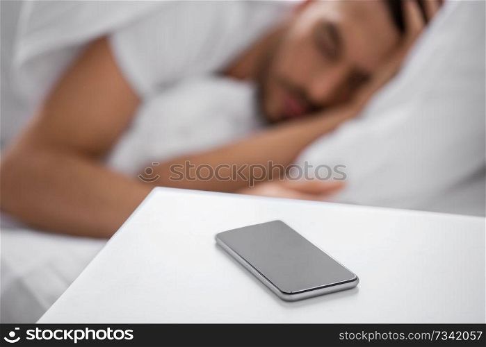 technology and people concept - close up of smartphone on bedside table near young man sleeping in bed at home in morning. smartphone on bedside table near sleeping man