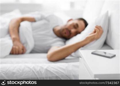 technology and people concept - close up of smartphone on bedside table near young man sleeping in bed at home in morning. smartphone on bedside table near sleeping man