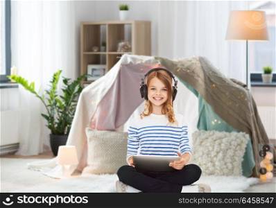 technology and people concept - girl with headphones and tablet pc listening to music at home over kids room and tepee background. girl with headphones and tablet pc at home