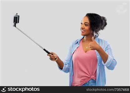 technology and people concept - happy african american woman taking picture by smartphone on selfie stick and showing thumbs up over grey background. african american woman taking selfie by smartphone