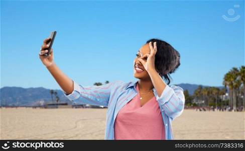 technology and people concept - happy african american woman taking selfie by smartphone over venice beach background in california. african american woman taking selfie by smartphone