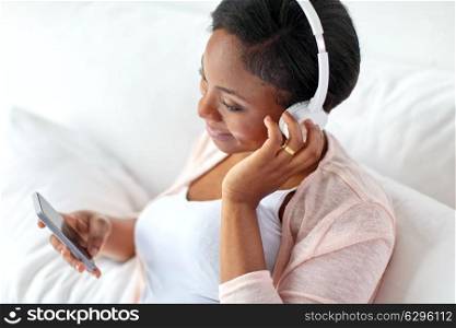 technology and people concept - happy african american woman with headphones and smartphone in bed at home. woman in headphones with smartphone