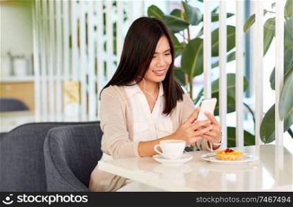 technology and people concept - happy asian woman with smartphone, earphones and cake listening to music at cafe or coffee shop. asian woman with smartphone and earphones at cafe